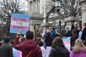 A person holds a sign with the transgender flag in front of the State House. (Brianna Silva/ File Photo)