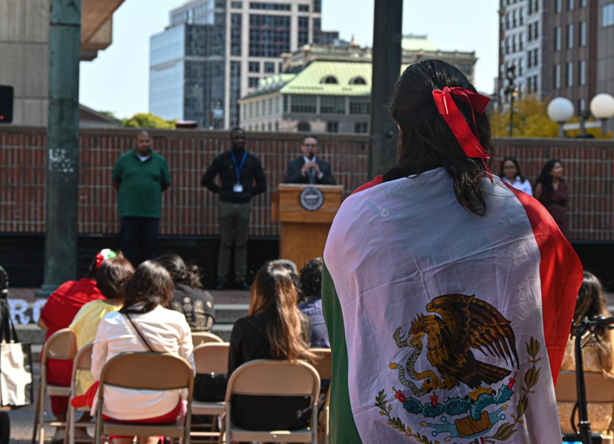 An onlooker draped in a Mexican flag watches as Felipe Cuéllar, the deputy consul of Mexico in Boston, gives a speech after the raising of the Mexican flag in City Hall Plaza on Mexican Independence Day on September 16, 2024 (Nick Peace / For the Beacon)